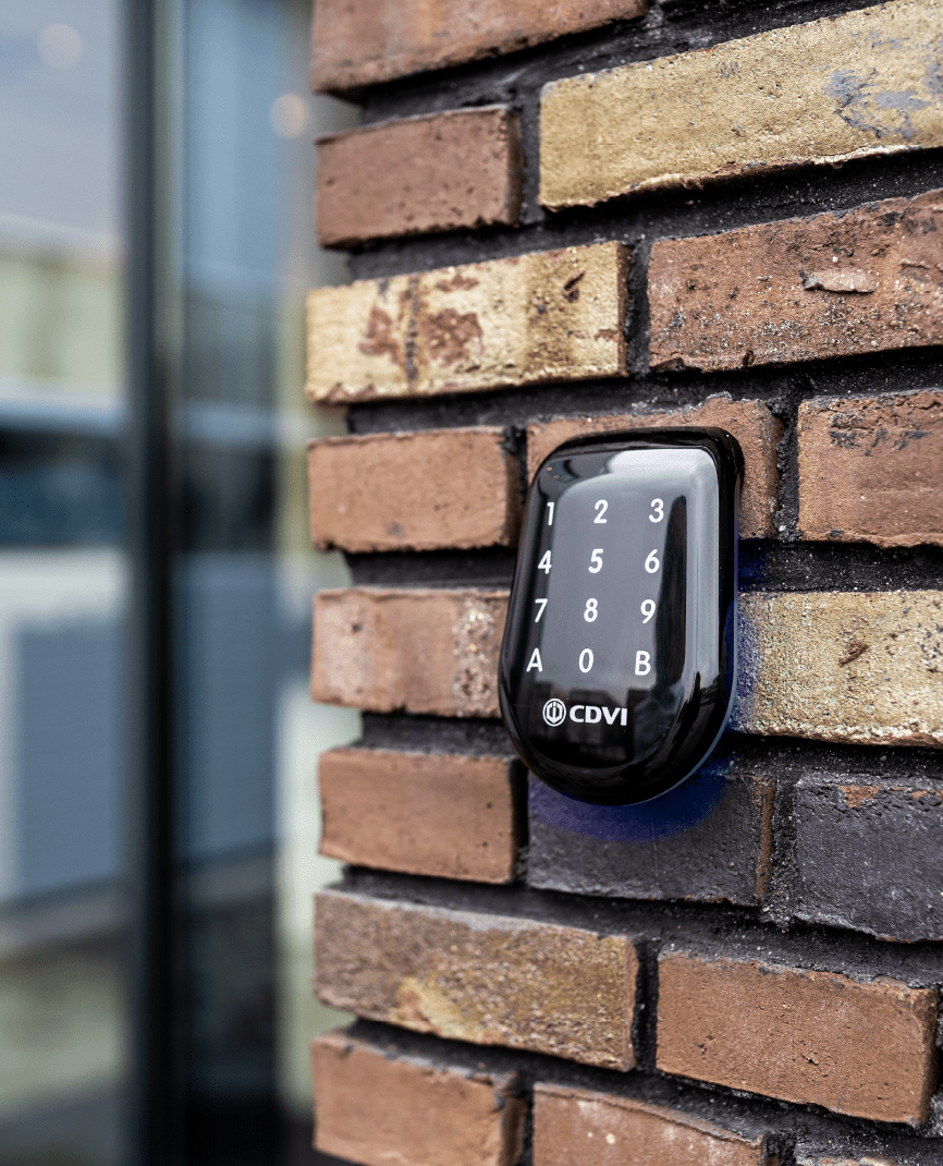 A black CDVI keypad mounted on a brick wall outdoors, with soft blue lighting underneath the keypad